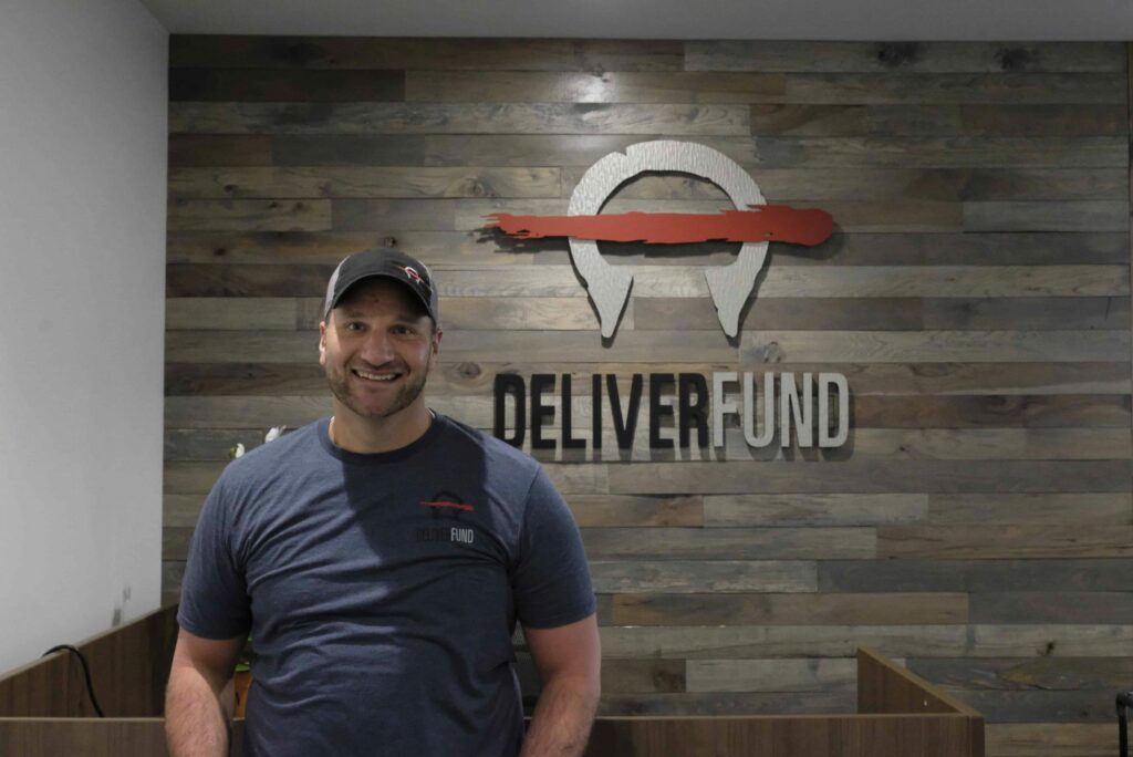 Shane Erickson standing in front of DeliverFund logo in HQ office